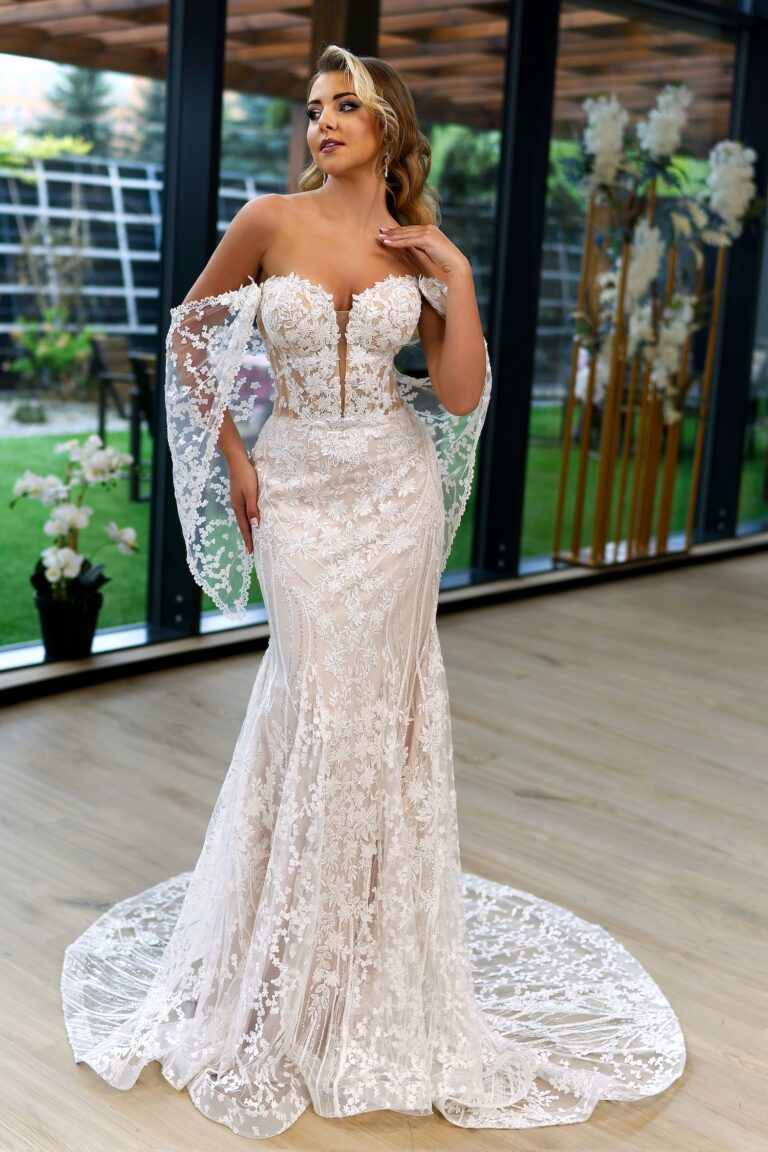 Eine Braut steht in einem modernen, lichtdurchfluteten Raum und trägt ein figurbetontes, weißes Brautkleid mit floralen Spitzenapplikationen. Das Kleid hat einen tiefen V-Ausschnitt und abnehmbare Spitzenärmel, die elegant herabfallen. Die Braut hat welliges, blondes Haar und berührt mit einer Hand sanft ihren Hals, während sie leicht zur Seite blickt. Im Hintergrund sind große Fenster und grüne Pflanzen zu sehen, die eine frische und natürliche Atmosphäre schaffen.